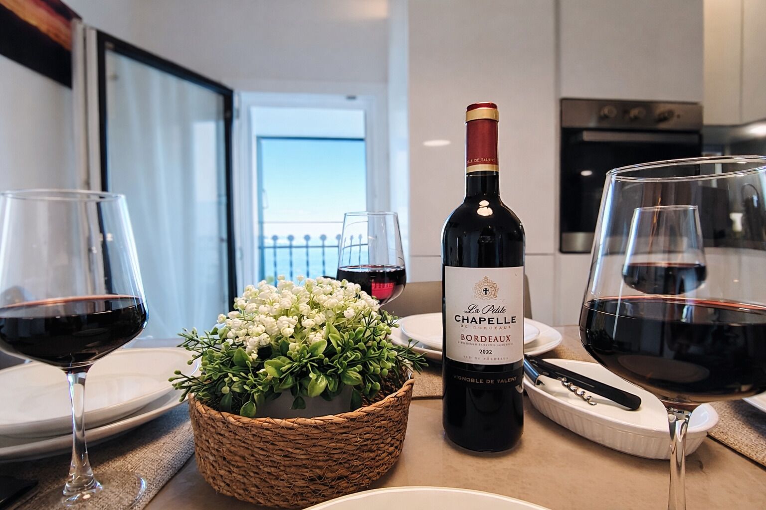 Une table à manger avec une bouteille de vin Chapelle Bordeaux 2032, deux verres de vin rouge, un panier tressé rempli de fleurs blanches et une assiette blanche.