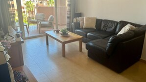 A living room with a black leather sofa, a wooden coffee table, and a potted plant. There is a sliding glass door leading to an outdoor area with a chair and a table. - **Introductory Discount** Gorgeous Apartment with Golf & Pool Views 14132 (Torre-Pacheco)
