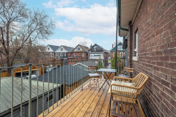 A wooden deck with a table and chairs, a metal railing, and a view of neighboring houses. - Bright + Stylish Apartment in High Park / Junction (Toronto)