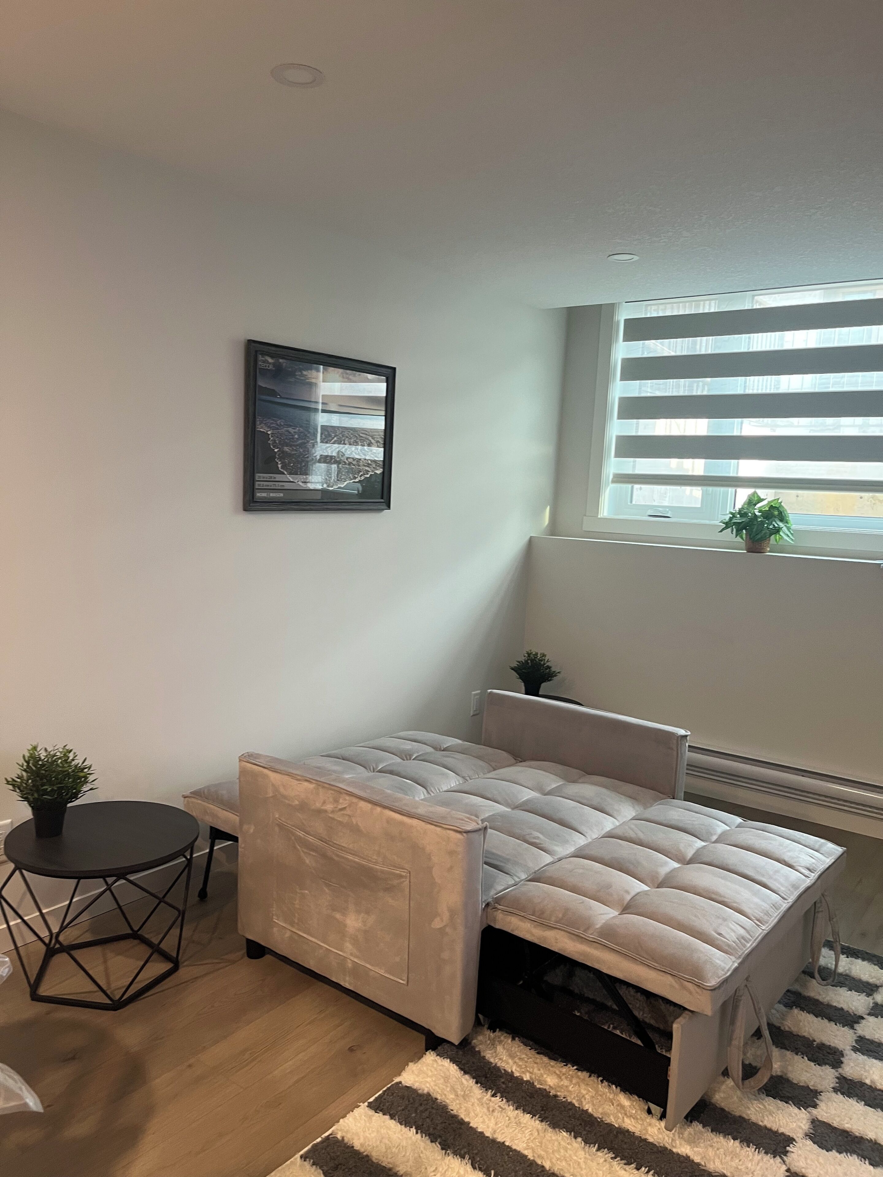A modern bedroom with a bed, a small round table, a potted plant, and a window with blinds.