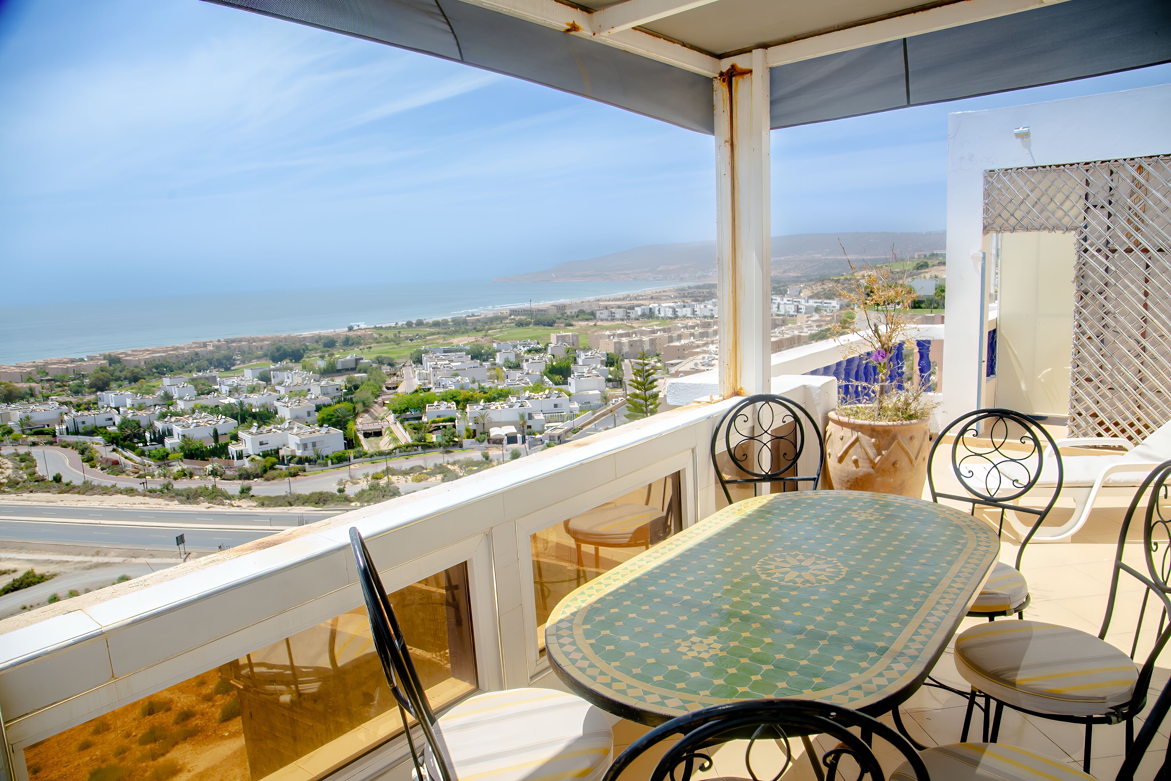 A balcony with a table and chairs, overlooking a coastal town and the sea.