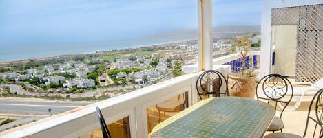 A balcony with a table and chairs, overlooking a coastal town and the sea.