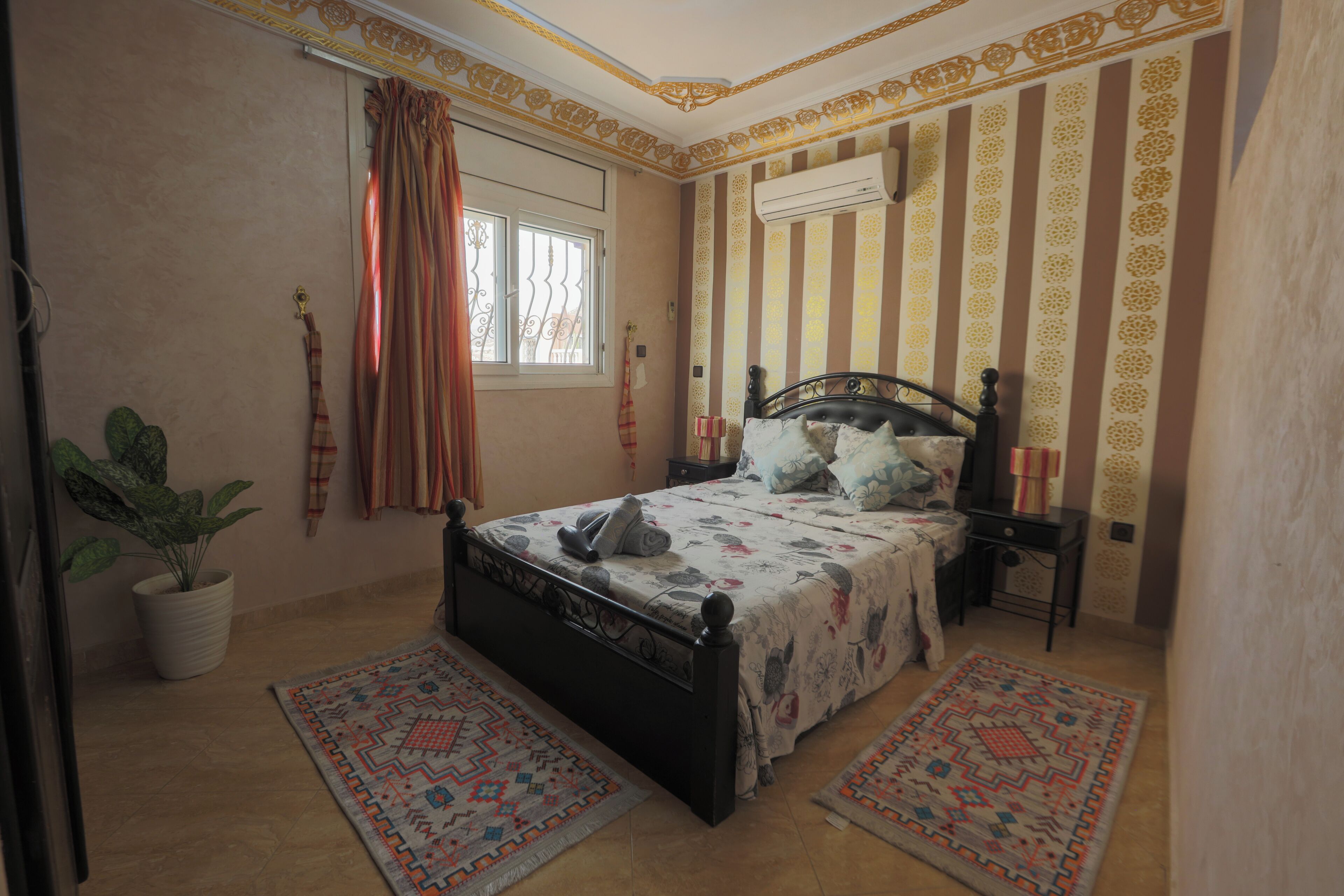 Une chambre à coucher avec un lit, une plante en pot, une fenêtre avec des rideaux, un appareil de climatisation et des murs à motifs.