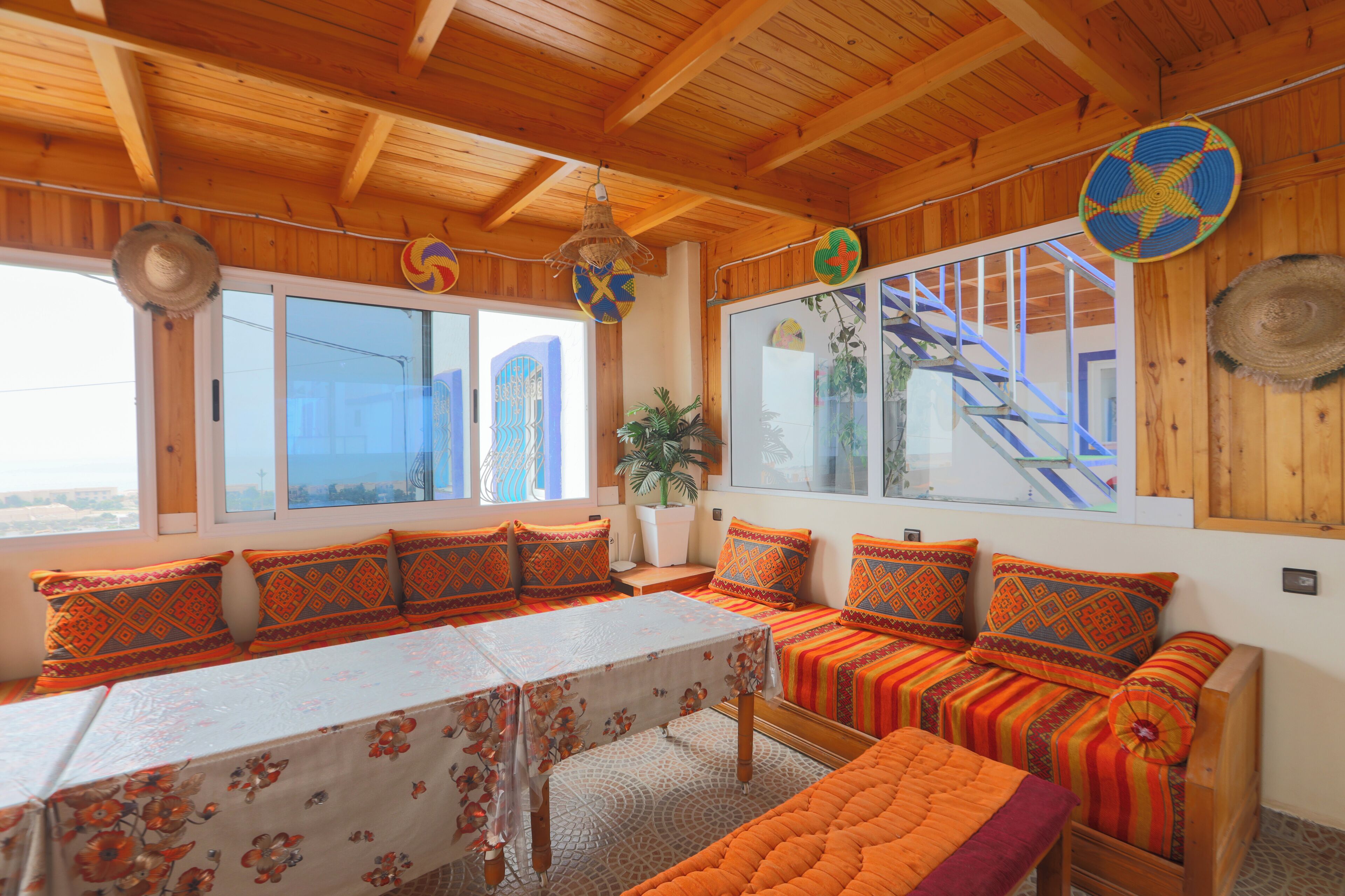 A room with wooden beams, a large window with a view, a table with a floral tablecloth, and a sofa with patterned cushions.