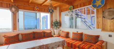 A room with wooden beams, a large window with a view, a table with a floral tablecloth, and a sofa with patterned cushions.
