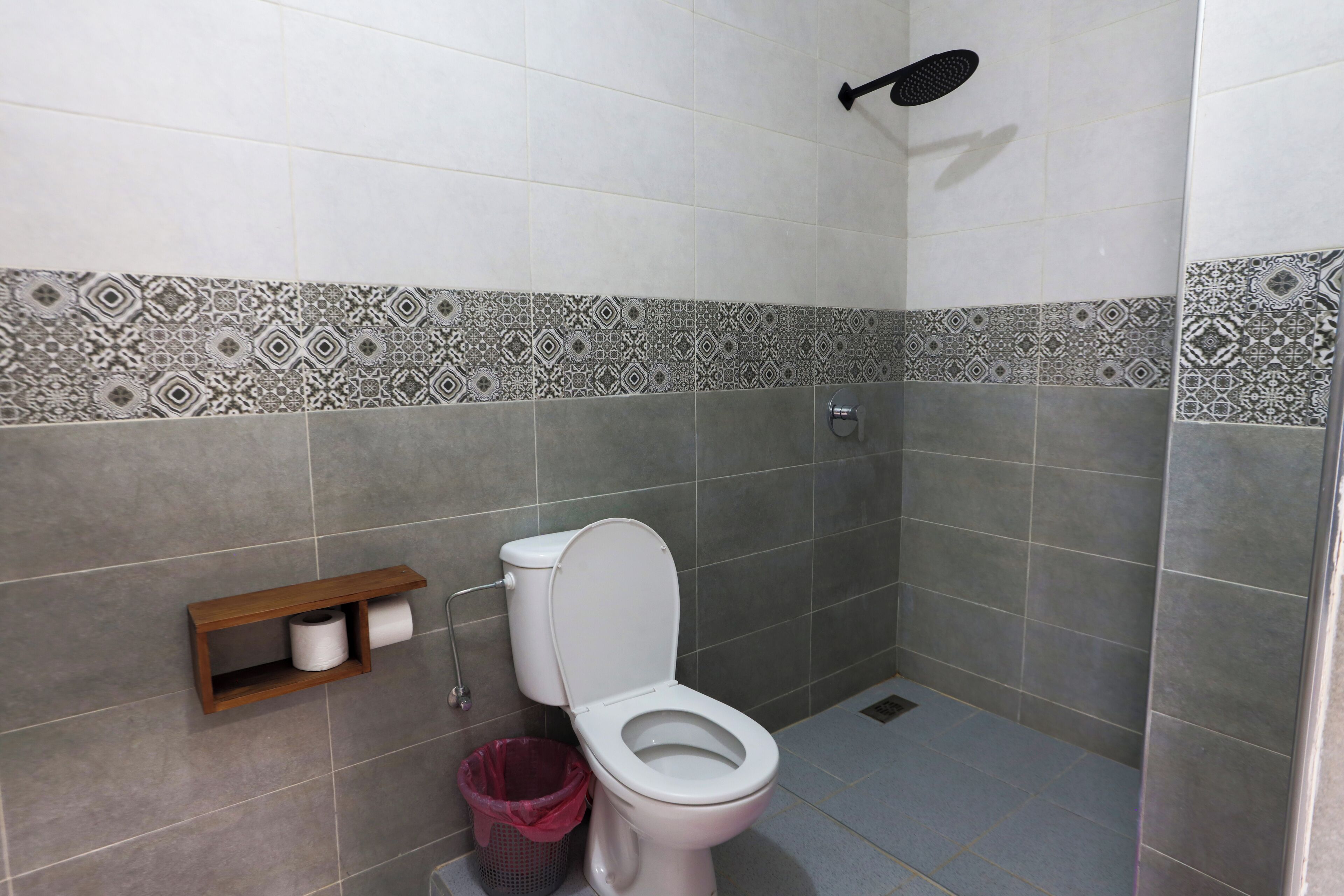 A bathroom with a toilet, a shelf with toilet paper, a trash bin, and patterned tiles on the walls.