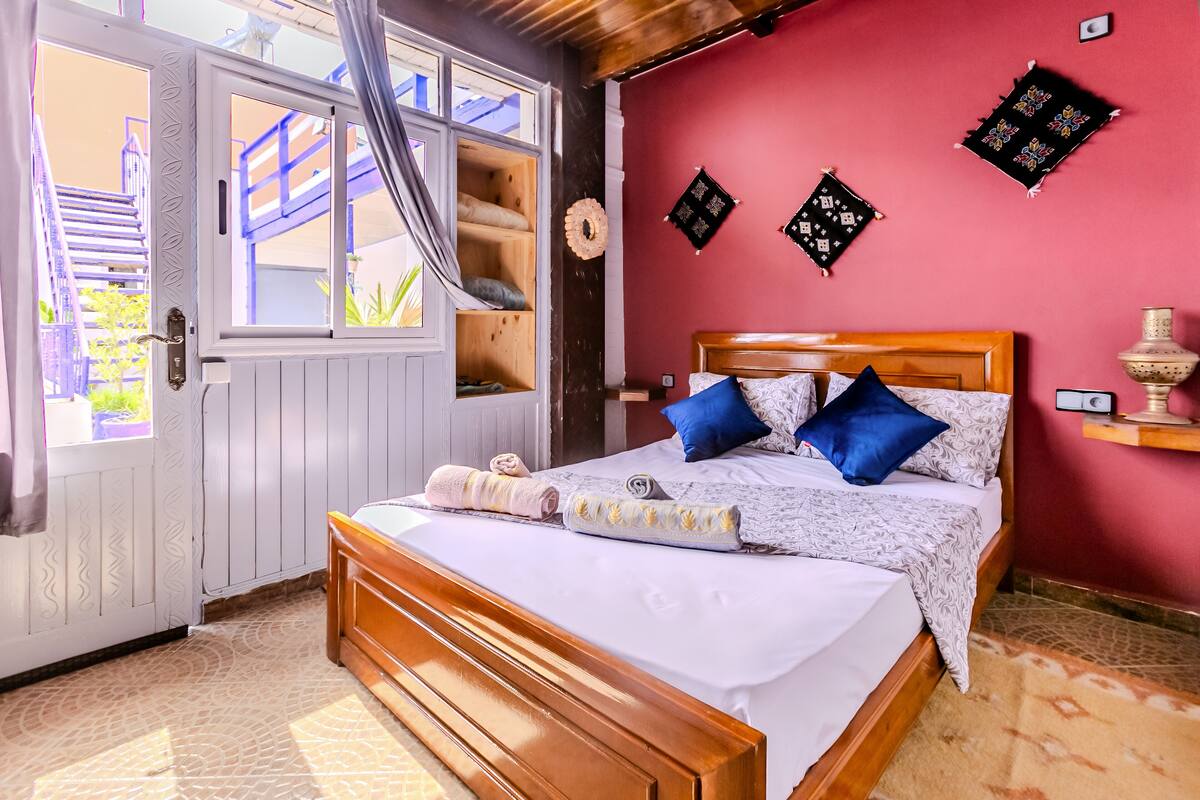 Un dormitorio con cama de madera, pared roja y ventana con vista a una escalera azul.