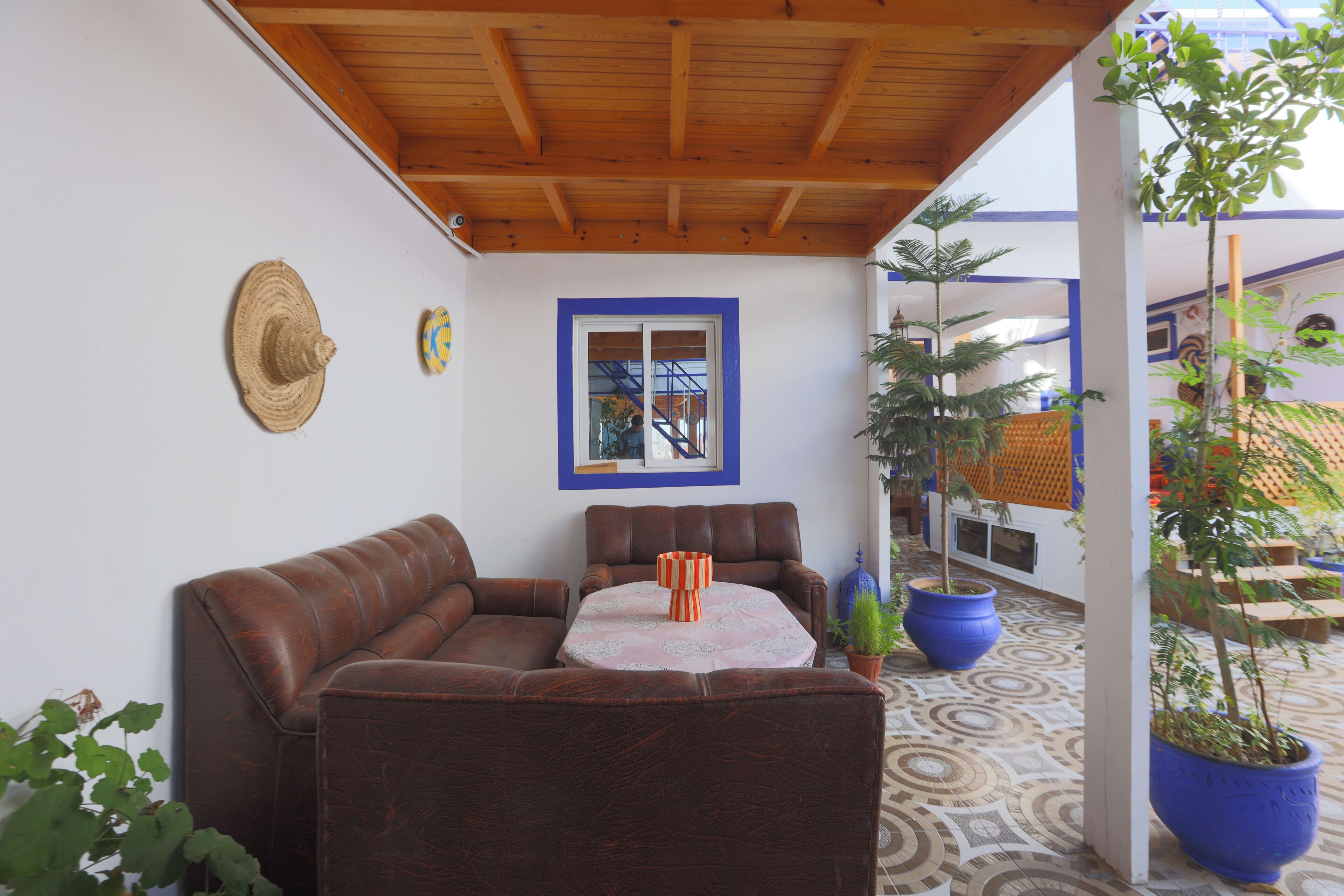 Een overdekt terras met een bruine leren bank, een ronde tafel, een houten plafond, een raam met blauwe omlijsting en potplanten.
