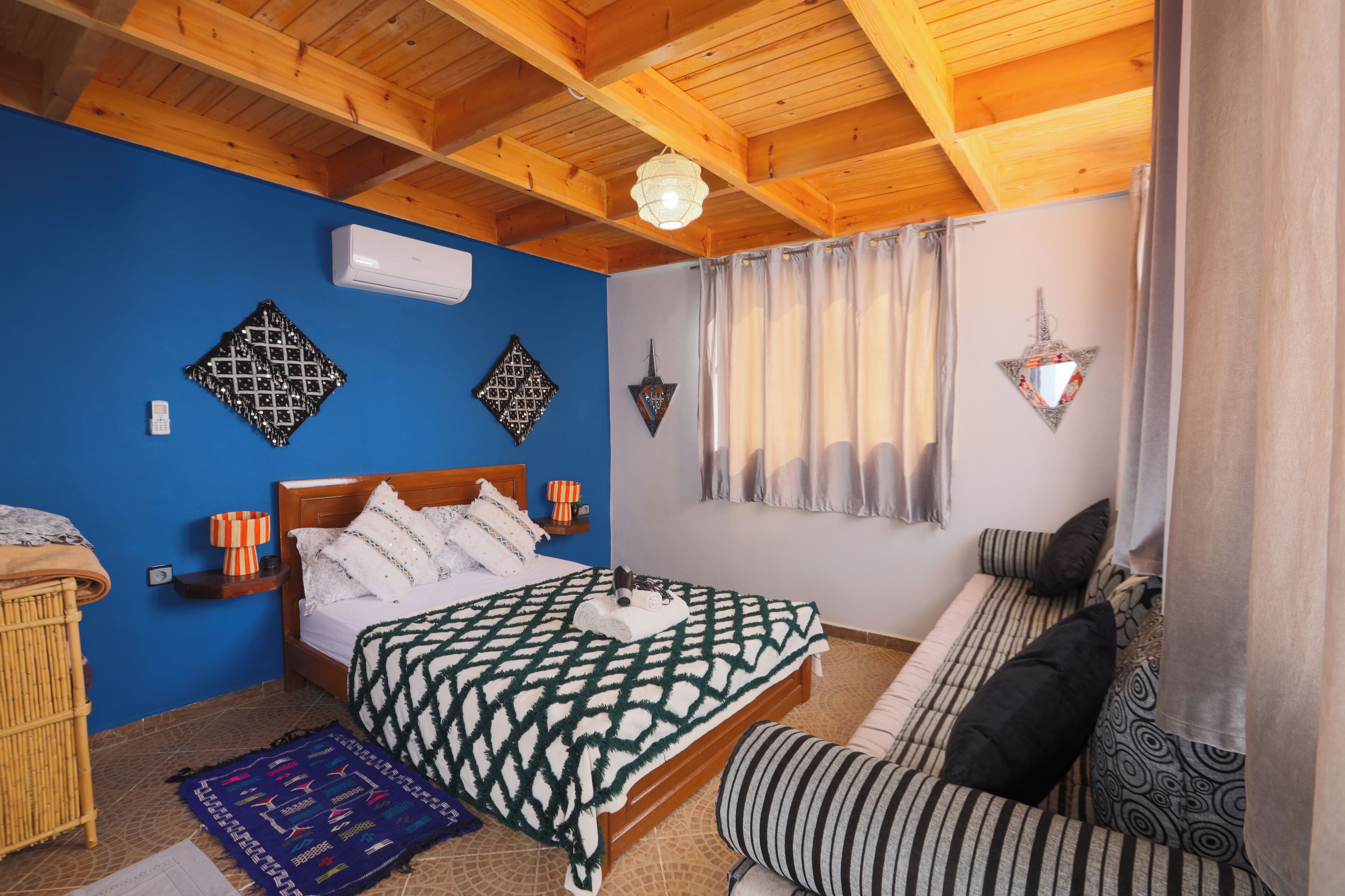 A bedroom with a wooden ceiling, a bed with a patterned bedspread, a sofa with cushions, a wall-mounted air conditioner, and decorative wall hangings.