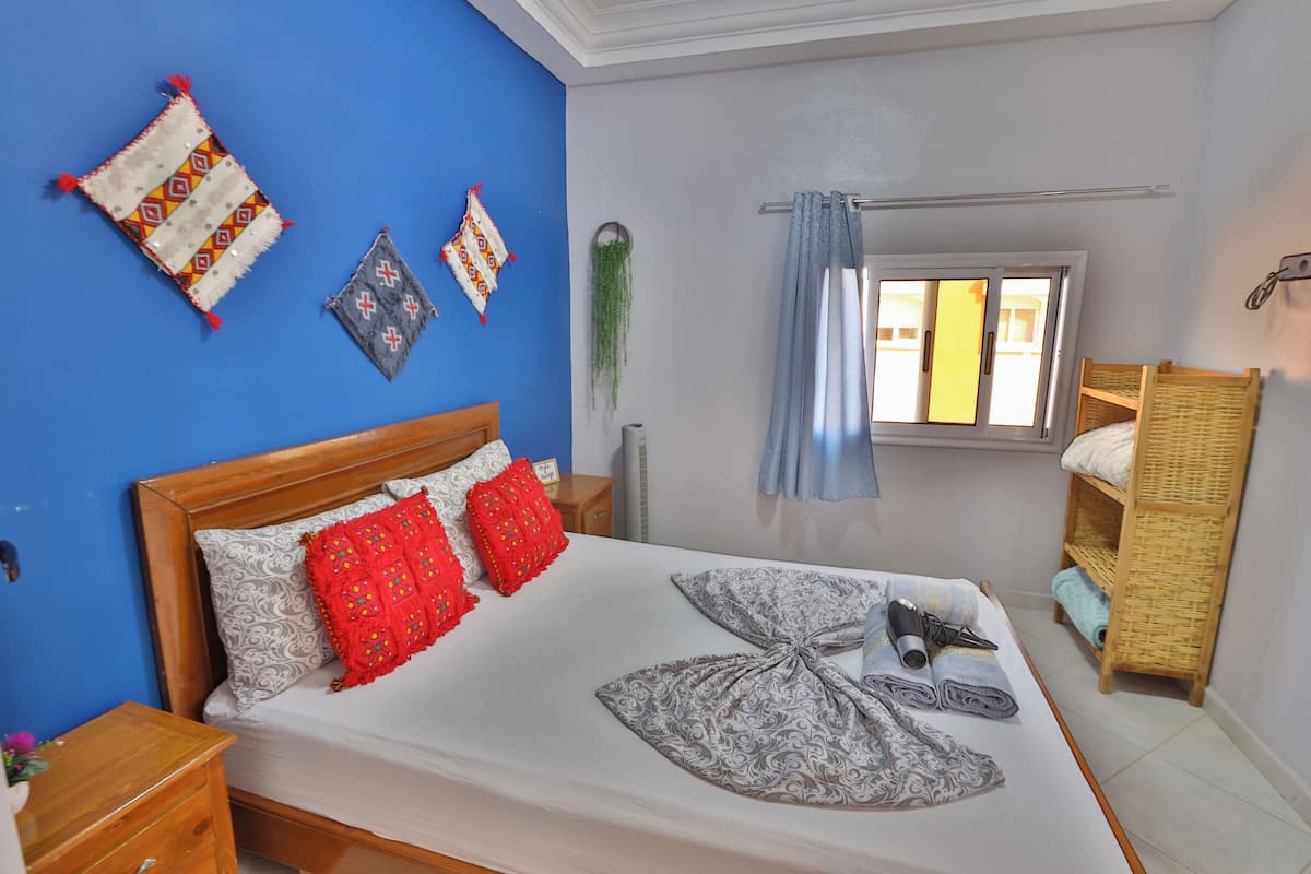 Un dormitorio con cama de madera, almohadas rojas y estampadas, una cortina azul y un ventanal pequeño.