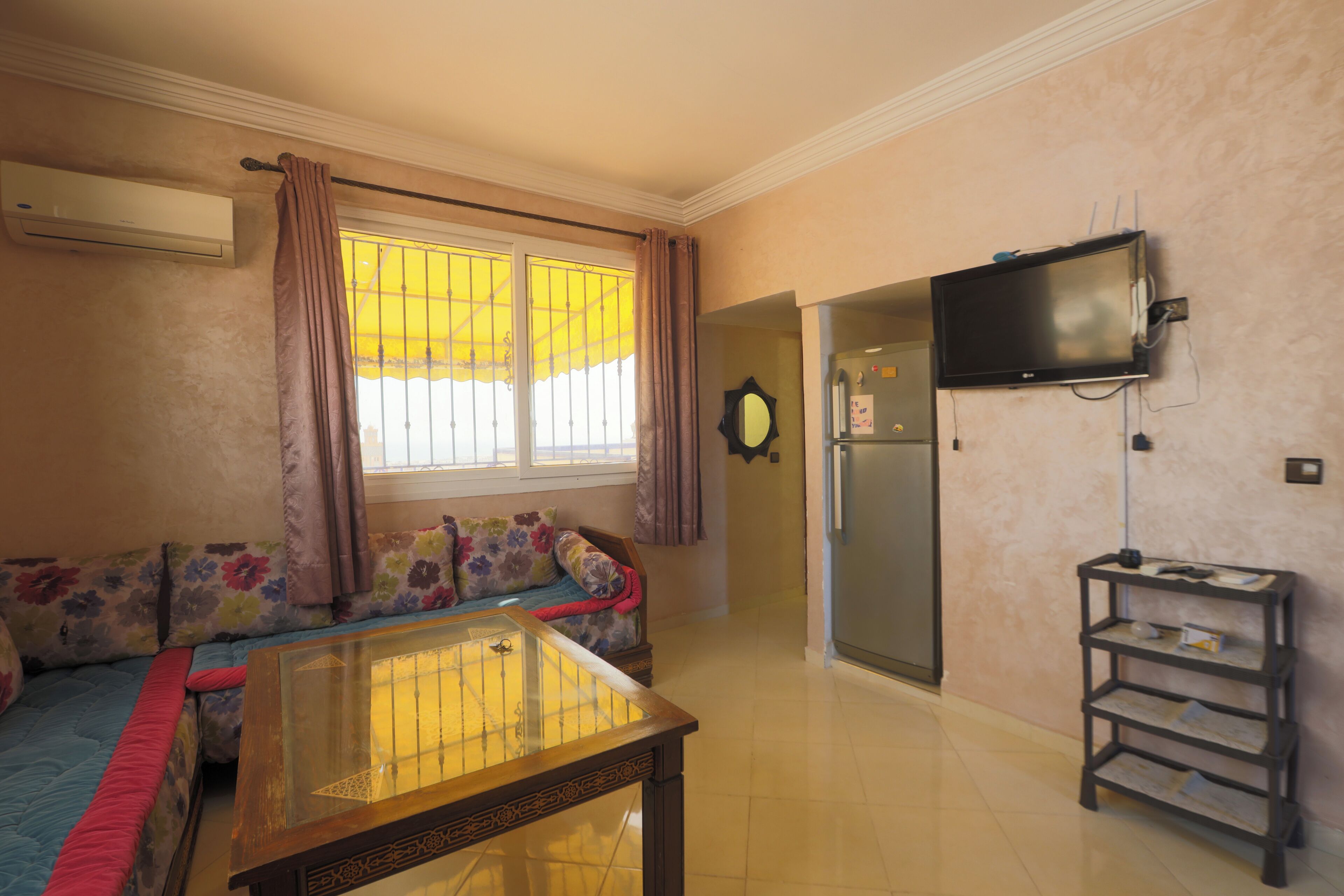 Una sala de estar con un sofá, una mesa de centro de vidrio, un televisor montado en la pared y un pequeño estante.
