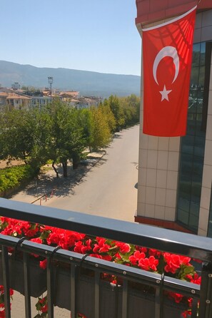 Ein Balkon mit Blick auf eine von Bäumen und Gebäuden gesäumte Straße, eine rote Flagge mit Halbmond und Stern sowie rote Blumen im Vordergrund.