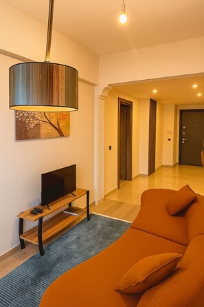 A modern living room with a flat-screen TV on a wooden stand, a large orange sofa, and a stylish pendant light. - Elegant Ephesus Suites - Artemis (İzmir)