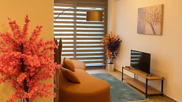 A modern living room with a curved sofa, a flat-screen TV on a stand, a large window with blinds, and a decorative tree.
