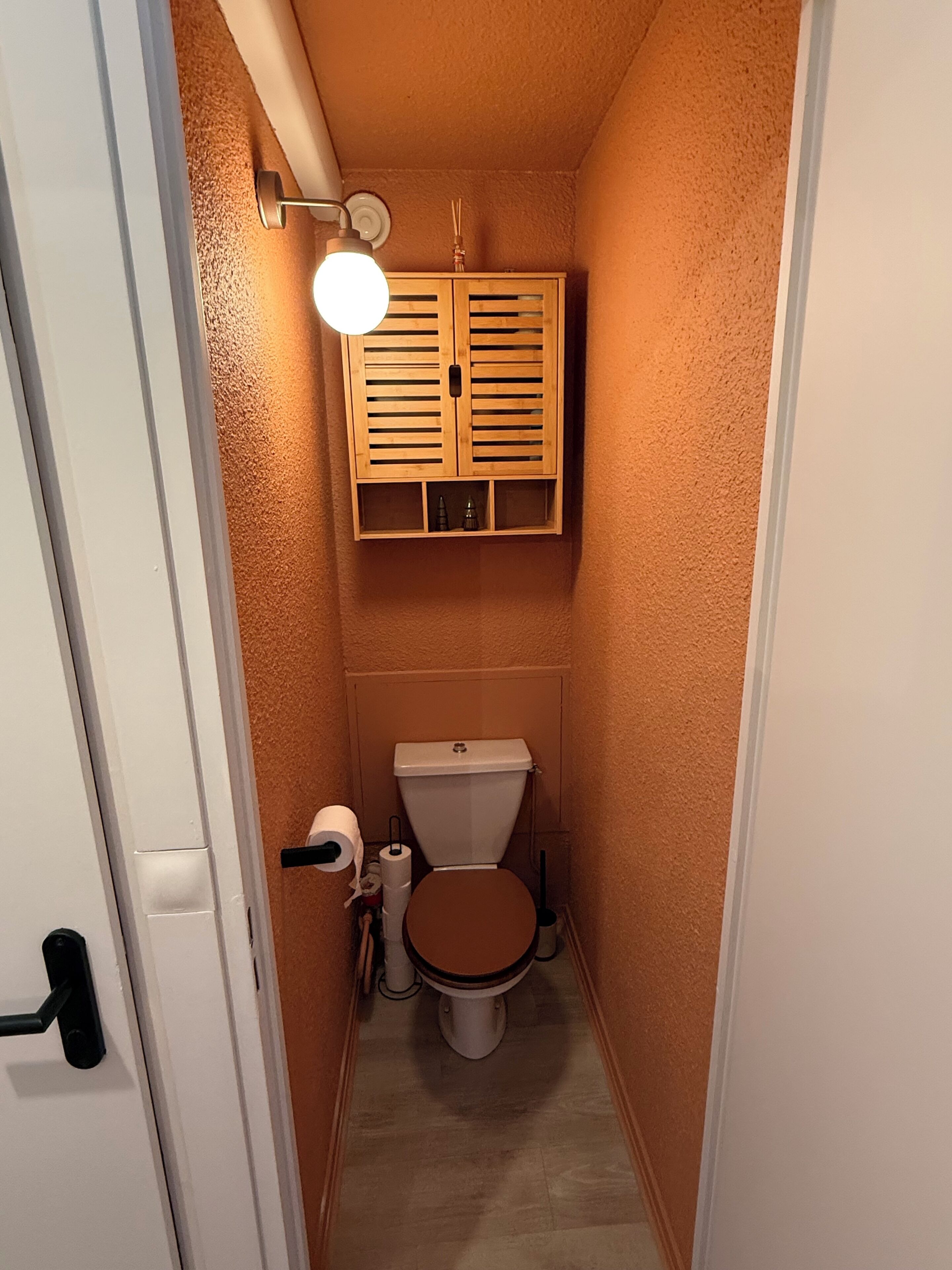 Een kleine badkamer met toilet, een houten kastje boven het toilet, en een wandlamp.