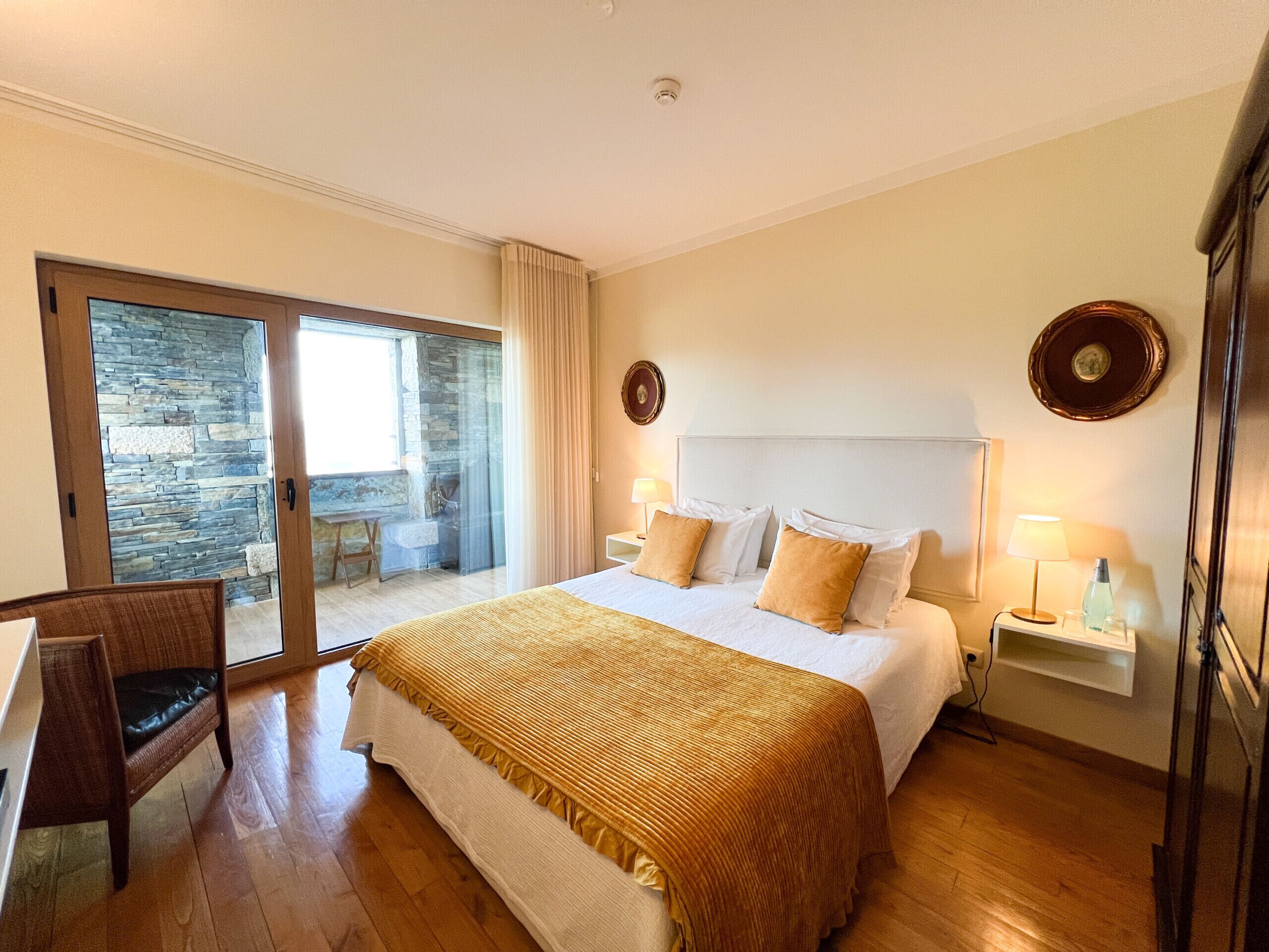 Une chambre d’hôtel avec un lit, un bureau, une chaise, une grande fenêtre et un mur en pierre.