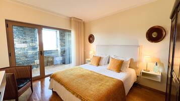 Une chambre d’hôtel avec un lit, un bureau, une chaise, une grande fenêtre et un mur en pierre.