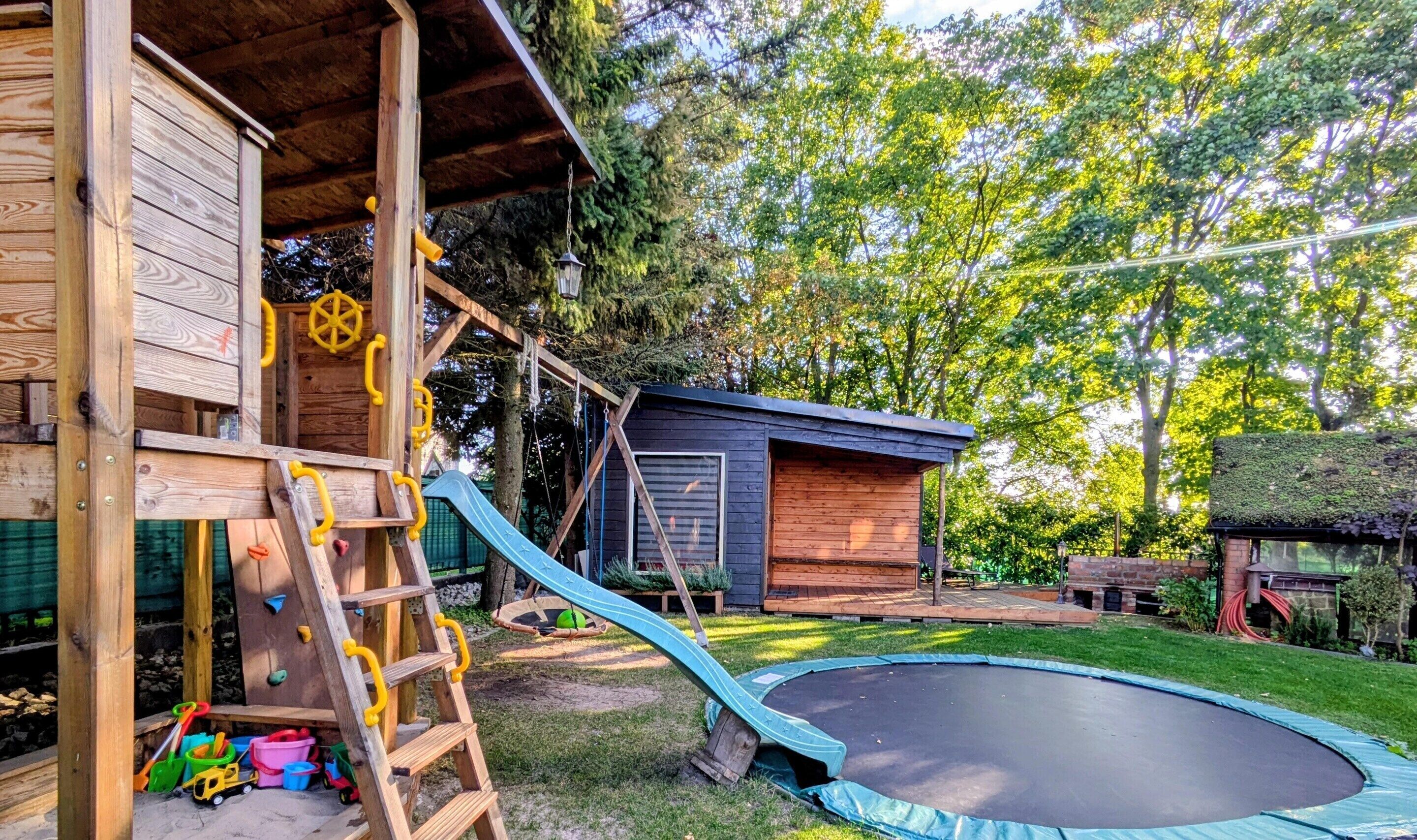 A wooden playhouse with a slide, a trampoline, and a small shed.