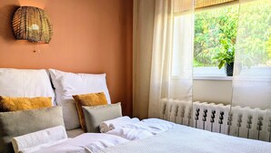 Une chambre à coucher avec un lit, des oreillers, une lampe fixée au mur, une fenêtre donnant sur un paysage verdoyant et un radiateur.