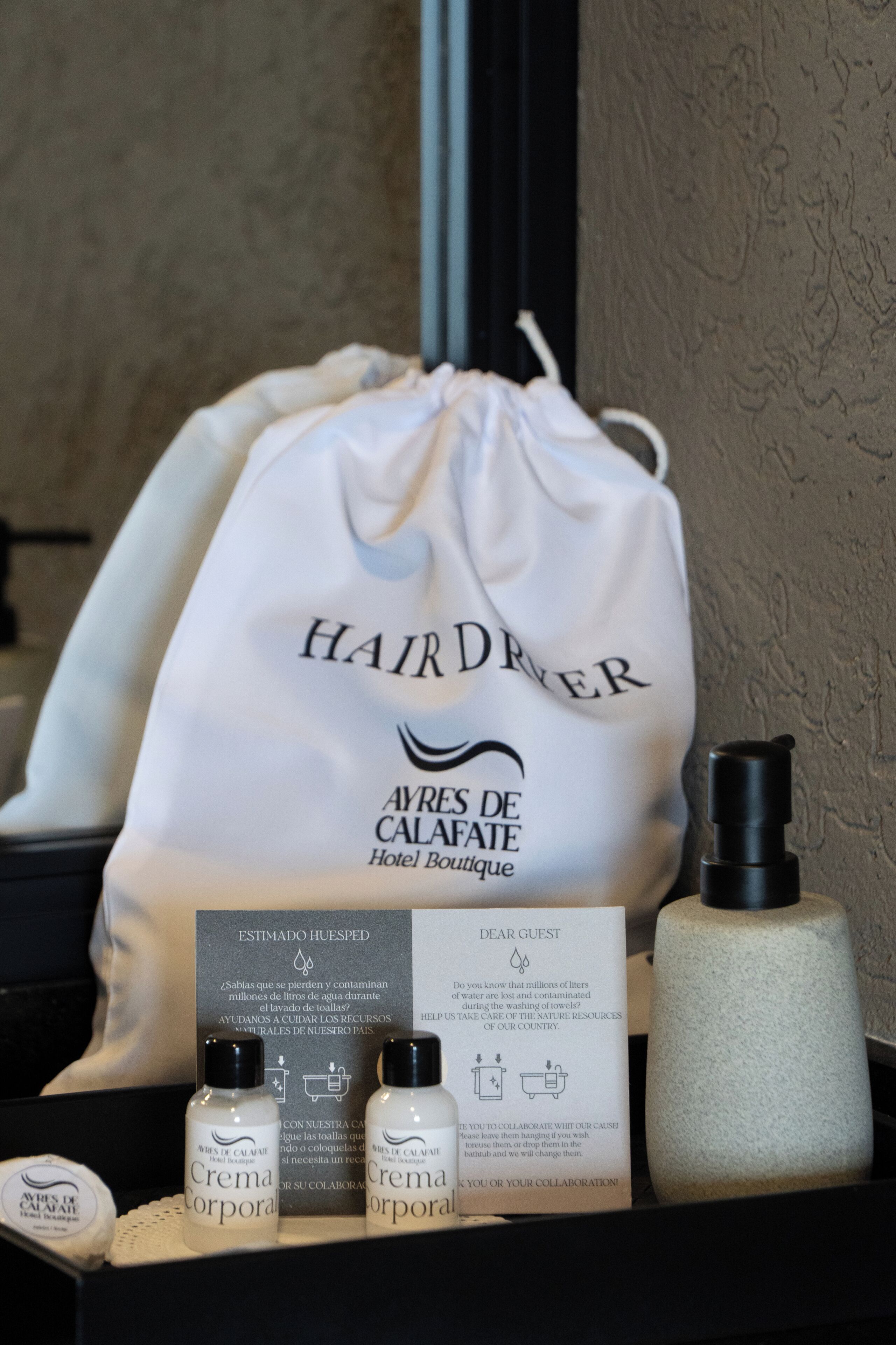 Un meuble-lavabo de salle de bain avec un sac blanc étiqueté « Sèche-cheveux », des flacons de « Crème pour le corps », un flacon noir et une serviette blanche.