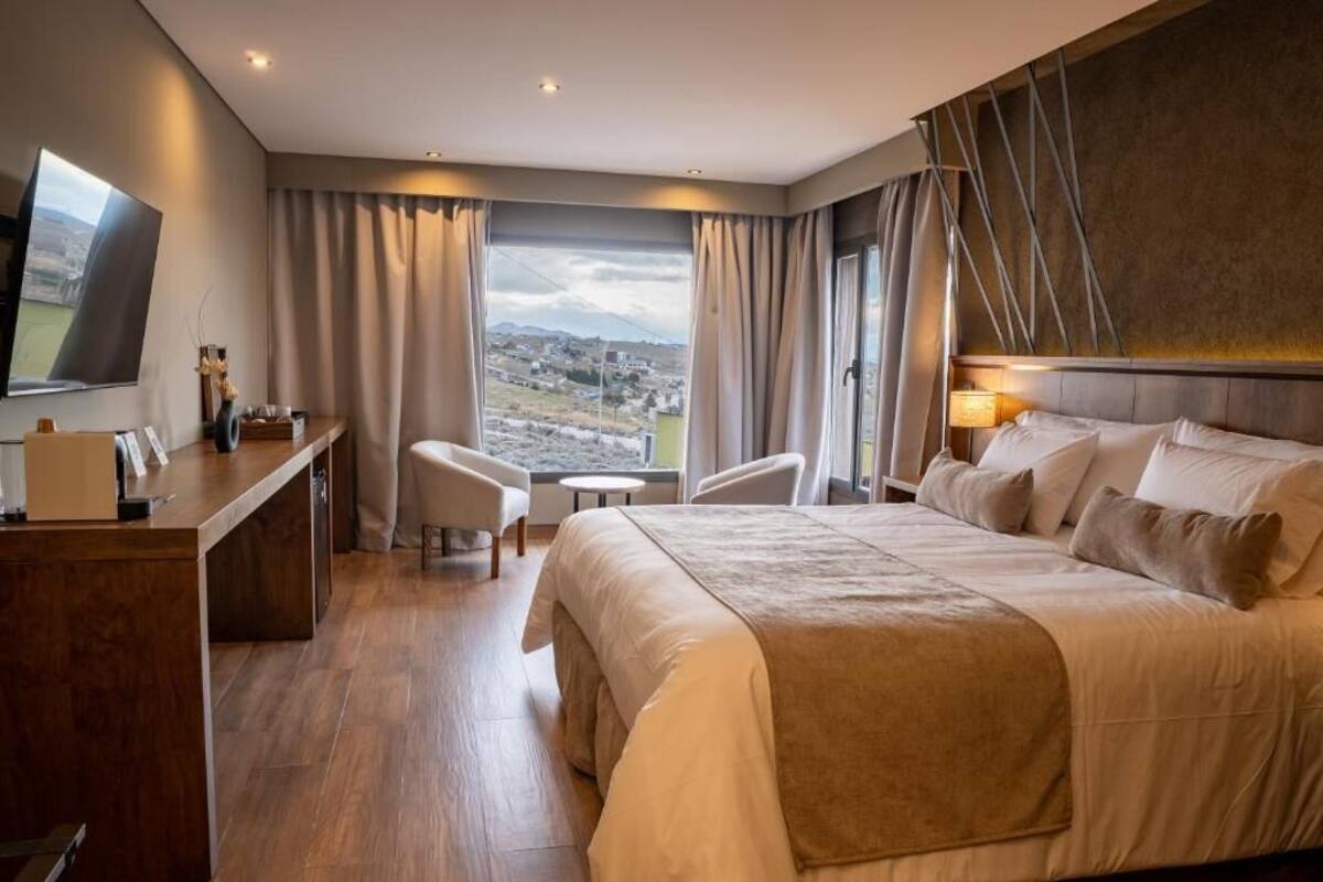 A hotel room with a large bed, a desk, a chair, a TV, and a view of a landscape.