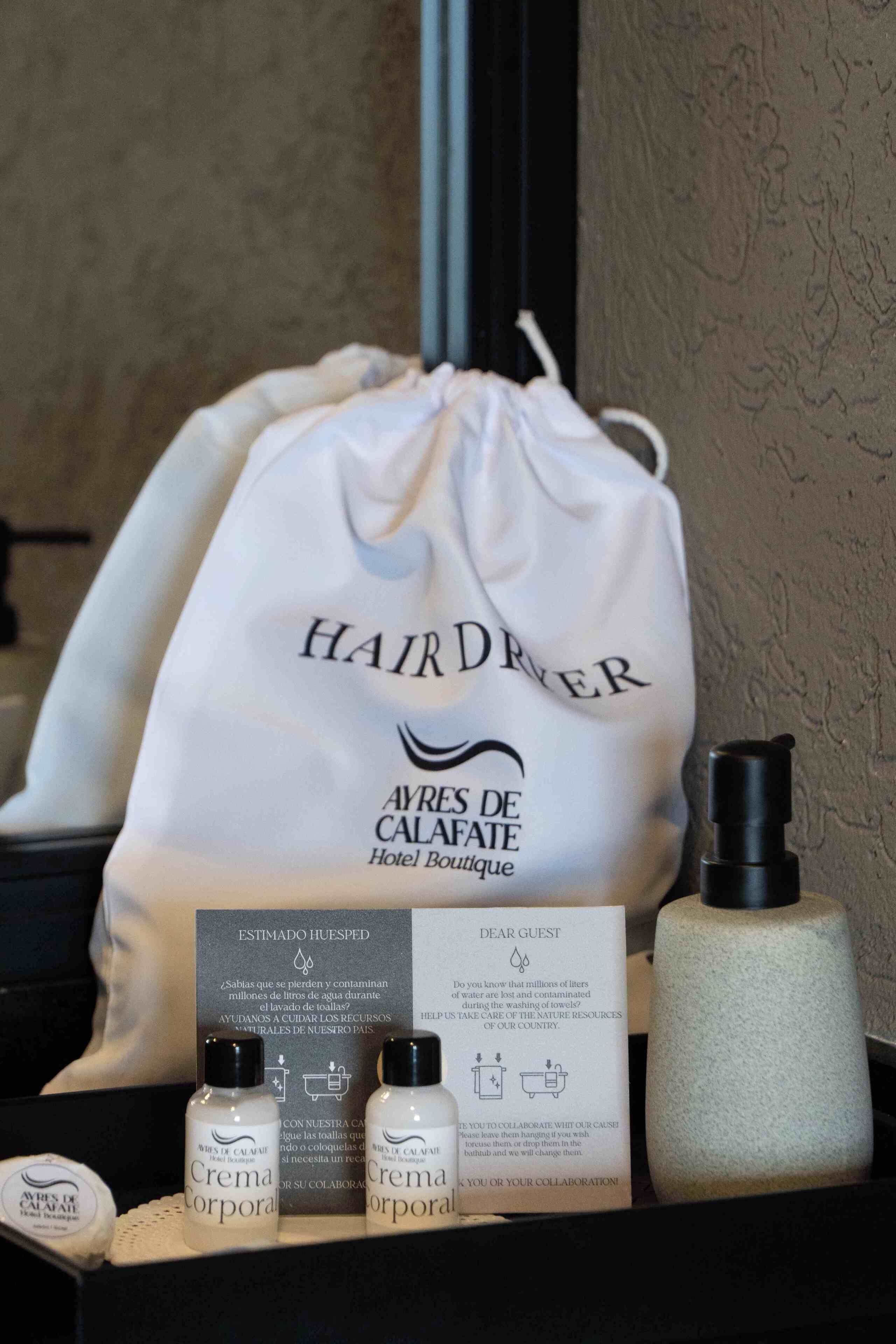 A bathroom setup with a white towel bag labeled 'HAIR DRYER,' a sign with water conservation message, and two bottles of 'Crema Corporal.'