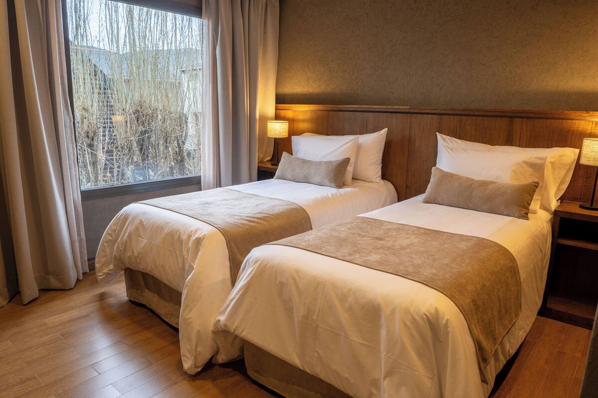 A hotel room with two beds, wooden headboard, bedside lamps, and a view of trees through the window.