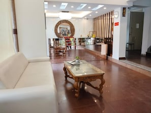 A lobby with a white sofa, a glass-top coffee table, and a reception desk with a mirror and plants. - Hotel Ambrosia by Zenith (New Delhi)