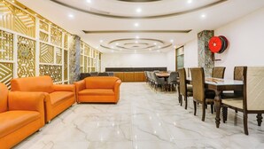 A spacious hotel lobby with a marble floor, orange sofas, a long dining table with chairs, and decorative wall panels.
