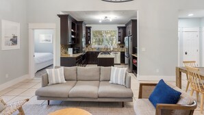 A modern living room with a sofa, a dining table, and a kitchen area visible through an open doorway. - Kemp’s Kove | 5BR Private Pool | <5 Min Walk to Beach | Sleeps 14 | Dog Friendly (Destin)