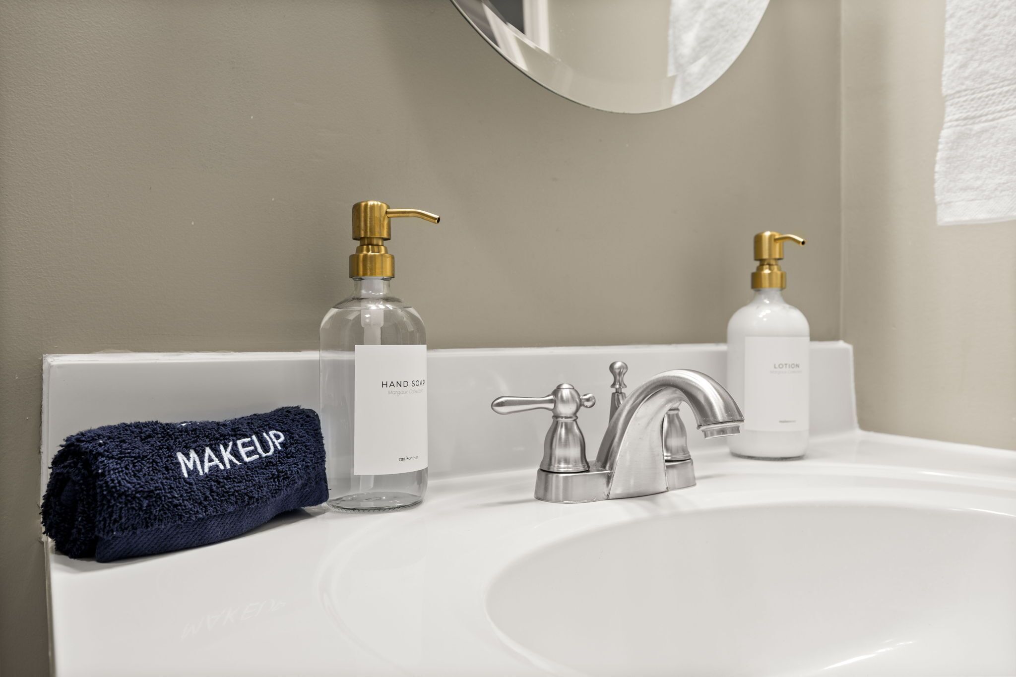Un baño con un lavamanos blanco, dos botellas de jabón para manos, una toalla con la etiqueta "MAKEUP" y un espejo sobre el lavamanos.