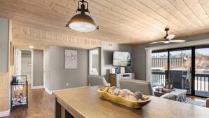 Amplia sala de estar con techo de madera, una gran mesa de comedor con un tazón de bocadillos, un televisor y vistas a un edificio a través de amplias ventanas.