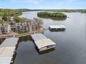 A waterfront hotel with multiple docked boats, a large building with balconies, and a small island with dense greenery. - Lakeside Condo Near Margaritaville w/ Dock, Boat Slip, Pool, & Mini Bar (Osage Beach)
