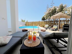 Uma área à beira da piscina com espreguiçadeiras, uma mesa com bebidas e um cercado de madeira.