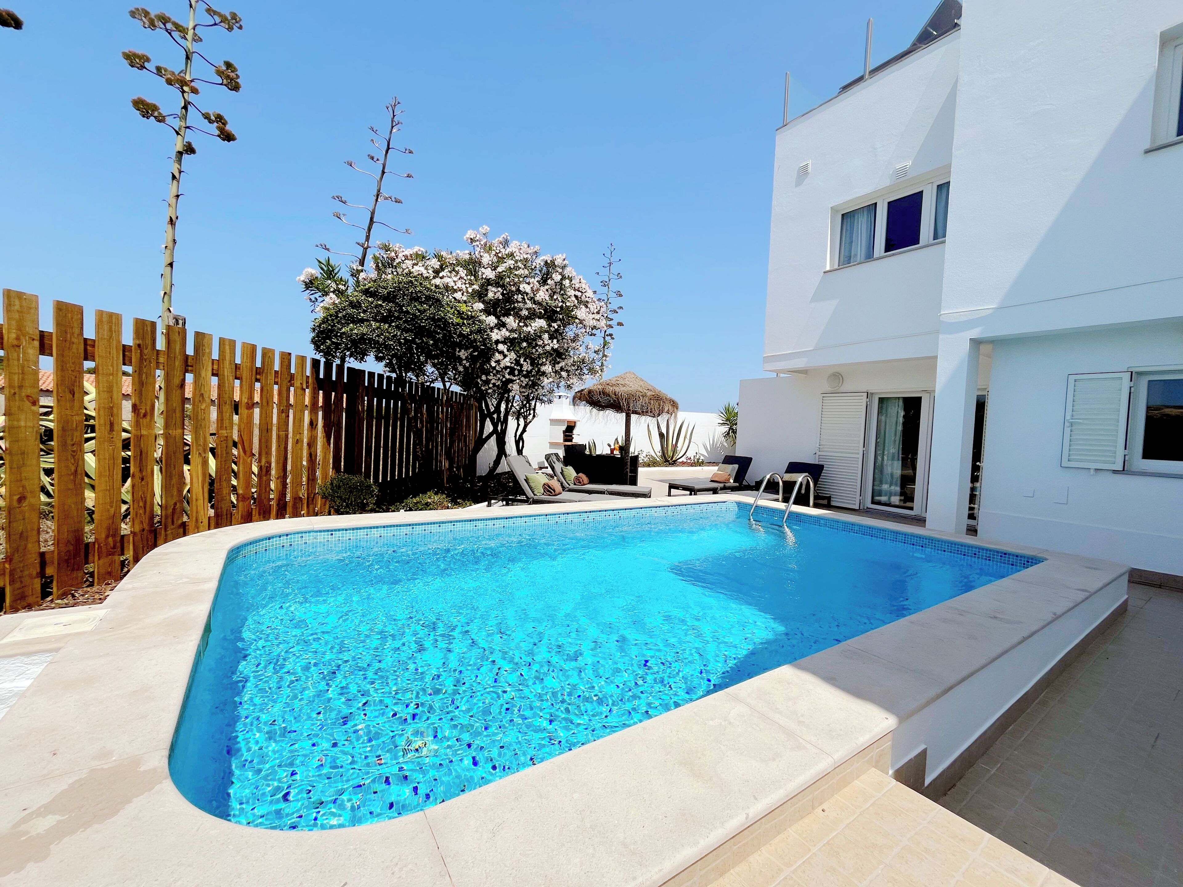 A rectangular swimming pool with a clear blue water surface, surrounded by a wooden fence and a white building with a balcony.
