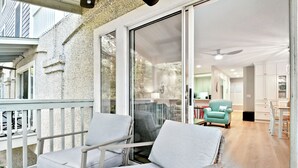 Ein Balkon mit zwei weißen Sitzgelegenheiten und einem Tisch, eine Glasschiebetür zu einem Küchenbereich mit weißen Schränken und einem türkisfarbenen Sessel.