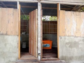 Una habitación con piso de concreto, paredes de madera y techo de metal. Hay una puerta de madera que conduce a otra habitación, y un contenedor naranja en la esquina.