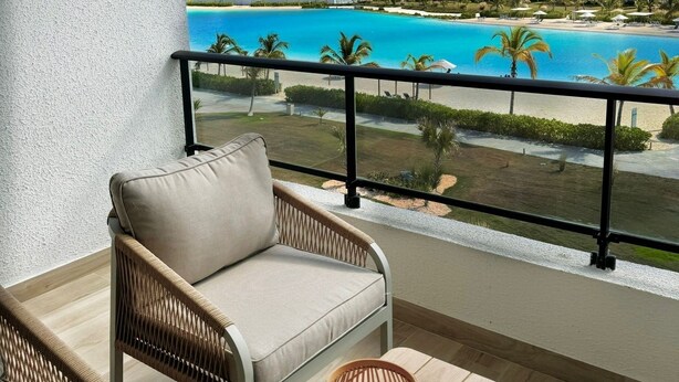 A balcony with wicker furniture, a view of a turquoise pool, and hotel buildings in the background. - Chic Boho Apartment Overlooking Crystal Lagoon (Punta Cana)