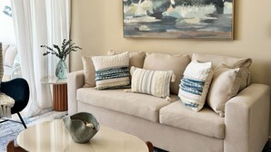A living room with a beige sofa, a round coffee table, and a painting of a seascape. - Chic Boho Apartment Overlooking Crystal Lagoon (Punta Cana)
