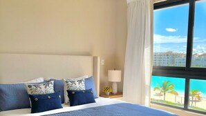 A large bed with a blue bedspread and matching pillows, a bedside table with a lamp, and a view of a beach through a large window. - Chic Boho Apartment Overlooking Crystal Lagoon (Punta Cana)