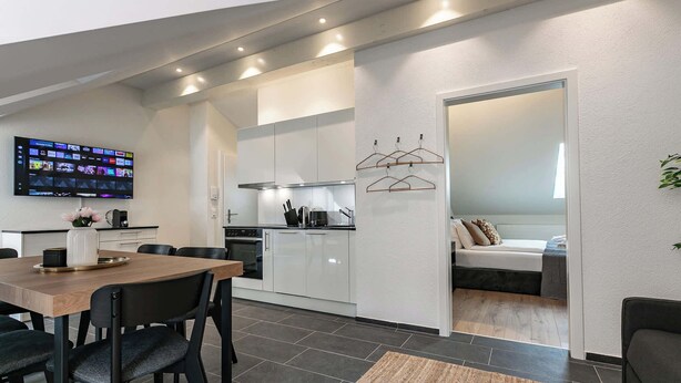 A modern kitchen with white cabinets, a dining area with a wooden table and black chairs, and a TV mounted on the wall displaying various channels. - book2stay 2.5-room apartment #8 Säntis up to 6 people (St. Gallen)