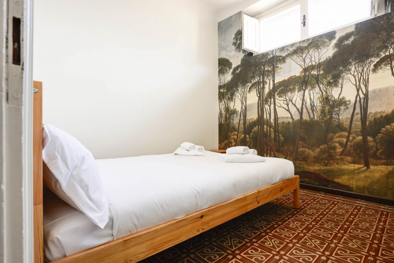 Un lit simple avec du linge de lit blanc et un cadre en bois, un sol à motifs et une grande fresque représentant une forêt sur le mur.