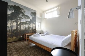 A bedroom with a bed, a painting of trees, a window, and a bedside lamp.