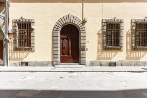 A building facade with a central arched entrance, two barred windows, and a stone-paved ground.