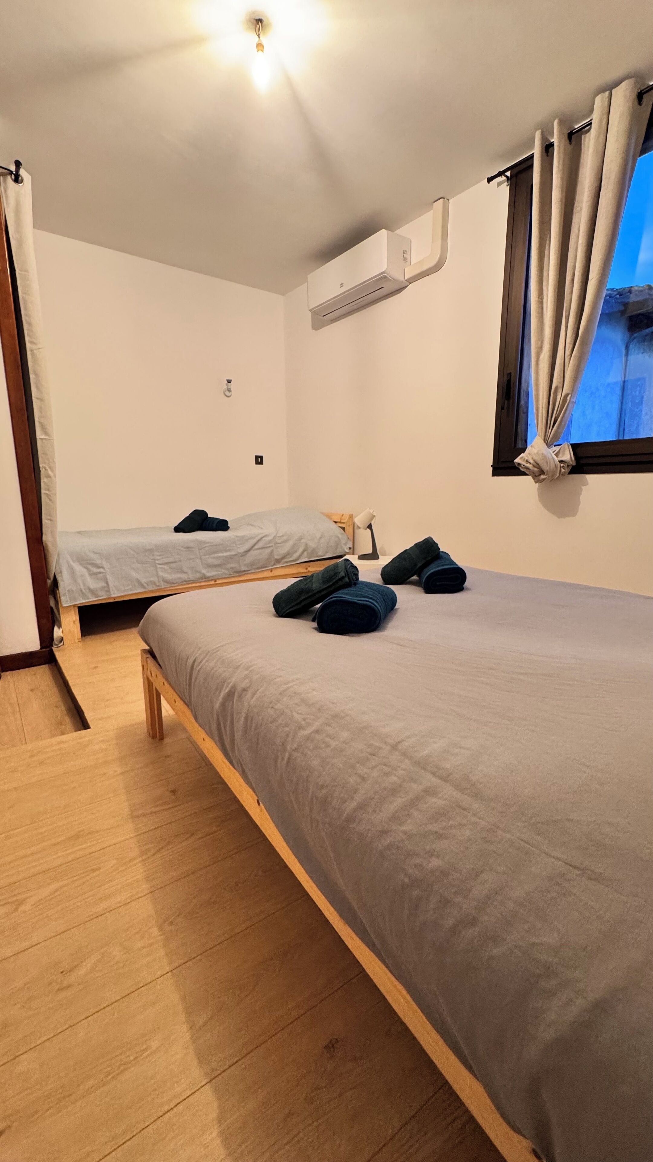 Un dormitorio con dos camas individuales, suelo de madera, unidad de aire acondicionado, ventana con cortinas y una lámpara de noche.