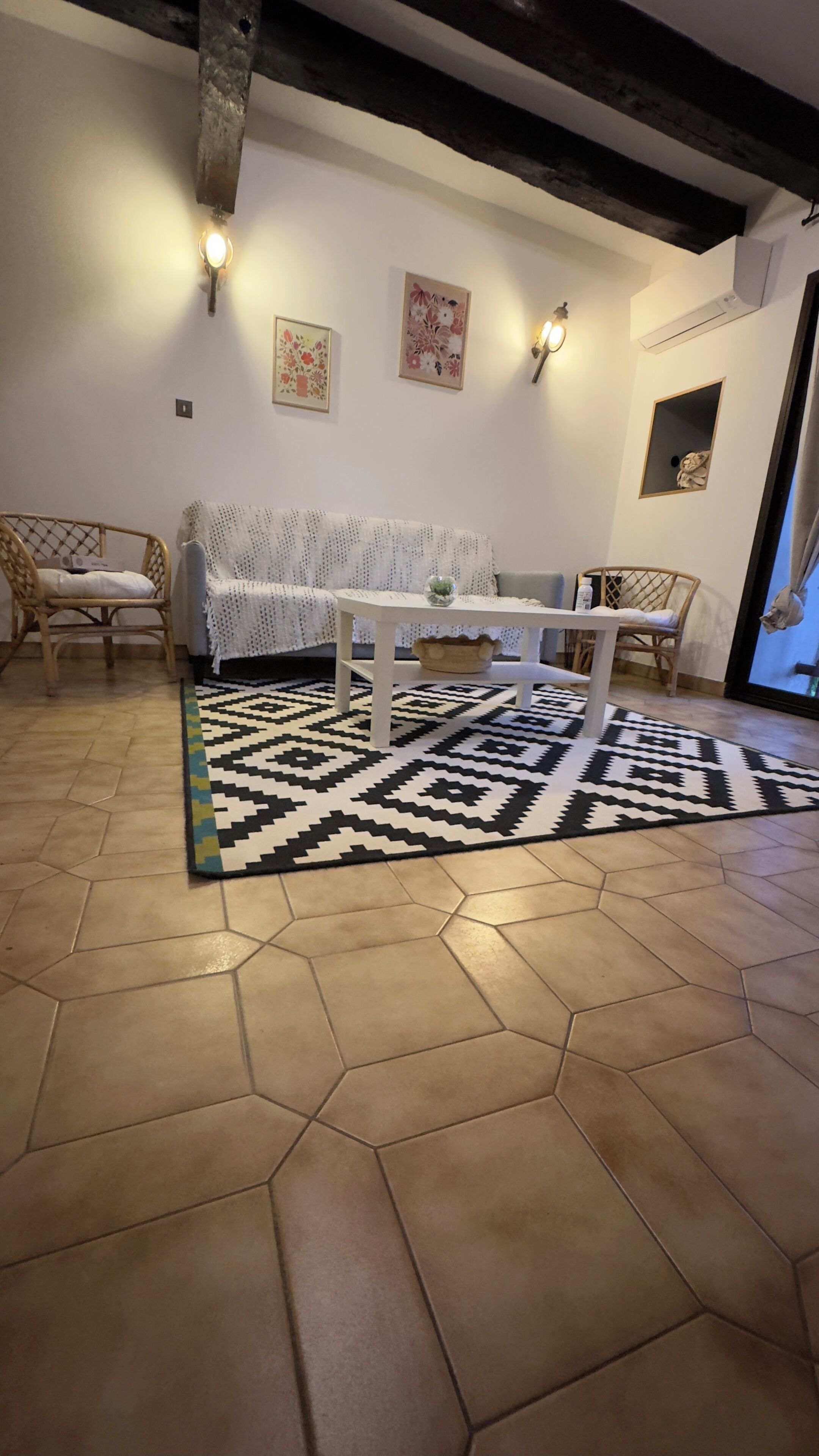 A living room with a hexagonal tiled floor, a white sofa, two wicker chairs, a white coffee table, and a patterned rug.