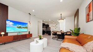 A modern living room with a flat-screen TV, a leather sofa, a dining area, and a kitchen in the background. - Luxury Oasis Near Disney 6Brds ,with Private Pool (Clermont)