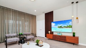 A modern living room with a large flat-screen TV, a sofa, a coffee table, and a dining area with a table and chairs. - Luxury Oasis Near Disney 6Brds ,with Private Pool (Clermont)