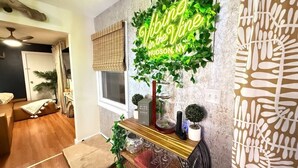 A living room with a wooden floor, a sofa, a coffee table, a wall-mounted neon sign, a decorative wall with a leaf pattern, and a small potted plant.