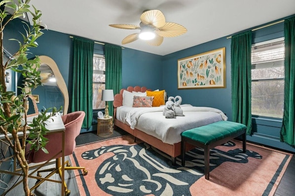 Une chambre à coucher comprenant un lit, un bureau, une chaise, une plante, un ventilateur de plafond et une fenêtre avec des rideaux.